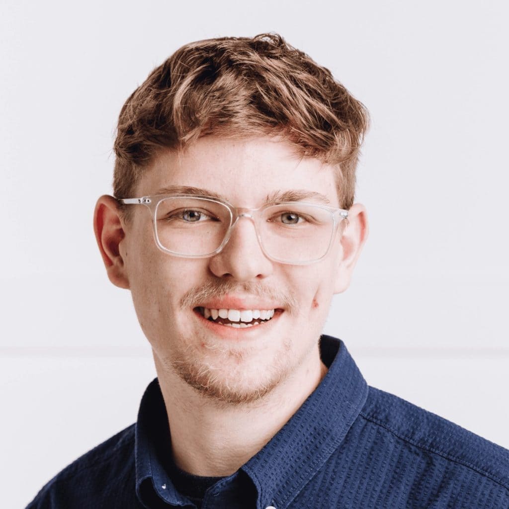 A smiling young man with brown hair and glasses wearing a blue collared shirt.