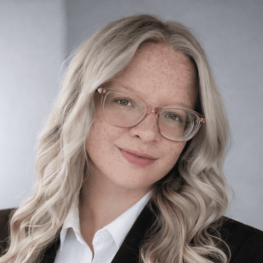 Portrait of a woman with long wavy blonde hair and glasses wearing a white shirt and dark jacket.