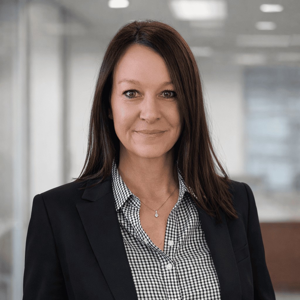 A smiling professional woman with long brown hair wearing a black blazer and checkered shirt in an office.